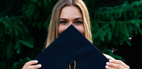Tjej som tagit examen i USA står med sin hatt och håller den framför delar av ansiktet.
