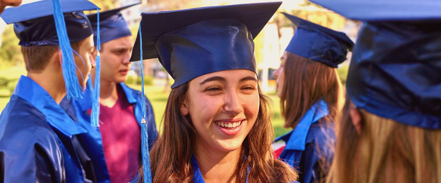 En glad high school student ler stort på sin examensdag, i en grupp med flera. De har på sig blå kappa och hatt med tofs.