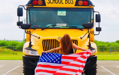En student står framför en gul skolbuss och håller en stor amerikansk flagga