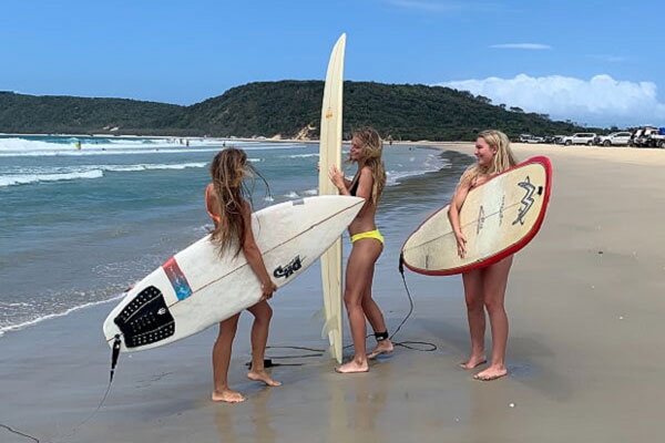 Tre tjejer med varsin surfbräda, står på stranden på väg till vattnet